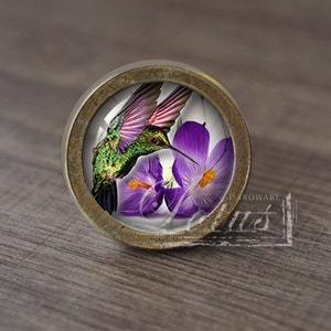 May include: A round, brass-colored cabinet knob with a glass insert featuring a green hummingbird hovering near purple crocuses.