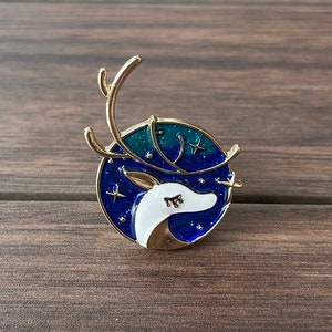 Puede incluir: Un pin de tono dorado con una silueta de cabeza de ciervo blanca sobre un fondo de cielo estrellado azul y verde.
