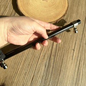 May include: A long, black metal object with silver detailing at each end. The item is held in a hand, set against a wooden background and a wooden slice.
