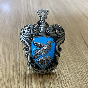 May include: A silver metal charm with a blue enamel crest featuring a raven and the words "Ravenclaw".