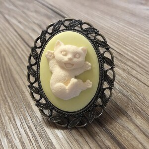Peut inclure: Une figurine de chat en céramique blanche dans un camée jaune avec un cadre orné argenté. Le chat est assis avec ses pattes levées et a une expression espiègle.