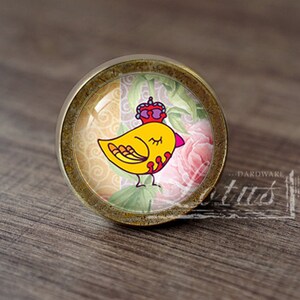Può includere: Un pomello rotondo per armadio dorato con un inserto di vetro che presenta un uccello giallo con una corona. L'uccello è circondato da un motivo floreale con rose rosa e foglie verdi.