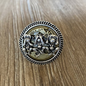 May include: A silver ring with a circular design featuring three monkeys covering their eyes, ears, and mouth. The monkeys are set against a gold background with a gear-like pattern.
