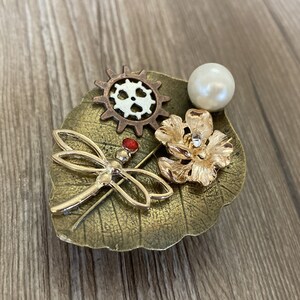 May include: A gold dragonfly brooch with a red gem on its body, a gold flower, a pearl, and a bronze gear on a bronze leaf.