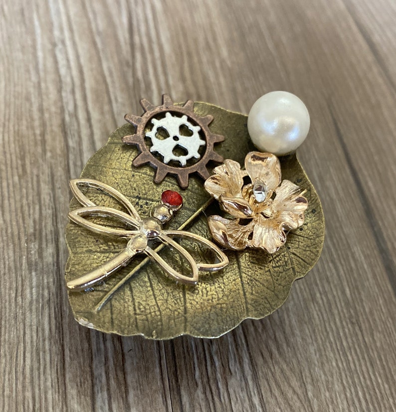 May include: A gold dragonfly brooch with a red gem on its body, a gold flower, a pearl, and a bronze gear on a bronze leaf.