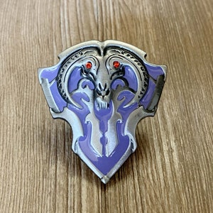 May include: A silver and purple enamel pin featuring a stylized skull with horns and red eyes. The pin is shaped like a shield.