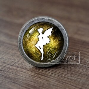 May include: A silver knob with a round, gold-colored glass insert featuring a white silhouette of a fairy with wings. The glass has a glittery texture.