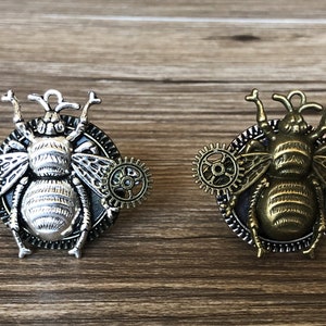 Może przedstawiać: Dwa broszki w stylu steampunk w kształcie pszczoły z misternymi detalami zębatek. Jeden broszka jest srebrny z czarnym tłem, a drugi jest złoty z czarnym tłem.