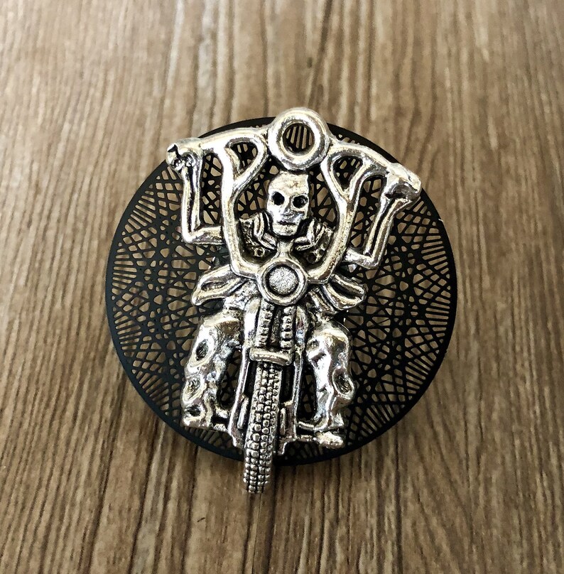 Op de afbeelding: Een zilverkleurige skeletmotorrijder-bedel met een zwarte filigraan achtergrond.
