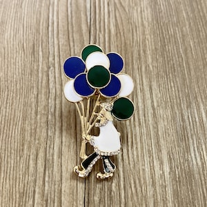 Puede incluir: Un broche de tono dorado con una figura de dibujos animados que sostiene globos azules, verdes y blancos. La figura lleva un vestido blanco y zapatos negros.