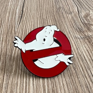 以下が含まれることがあります： ゴーストバスターズのロゴが描かれた赤と白のエナメルピン。赤い円の中に、驚いた表情をした白い幽霊が斜めの線で閉じ込められている。