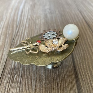 May include: A bronze leaf-shaped trinket box with a dragonfly, a flower, and a gear design. The box has a pearl-like knob on top.