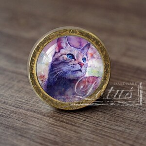 Może przedstawiać: Okrągły, mosiężny gałka do szafki z wkładką szklaną przedstawiającą akwarelę kota z niebieskimi oczami na tle fioletowych i różowych odcieni.