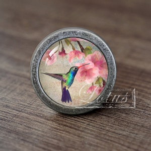 Op de afbeelding: Een ronde, zilverkleurige kastknop met een glazen inzetstuk met een kleurrijke kolibrie in vlucht, die zweeft in de buurt van roze bloemen.