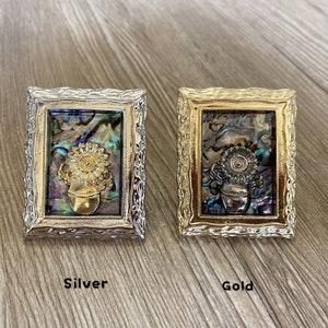 May include: Two small framed pictures with a gold and silver finish. The pictures feature a sunflower in a pot made of iridescent abalone shell. The frames are ornate and have a decorative border. The picture on the left is silver and the picture on the right is gold.