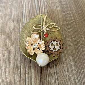 May include: A gold and bronze brooch featuring a dragonfly, a flower, a gear, and a pearl. The brooch is set on a leaf-shaped base.