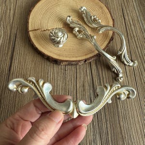 以下が含まれることがあります： シルバーカラーのキャビネット金具セット。装飾的なハンドル2つ、装飾的なノブ1つ、カーブしたプル1つが含まれています。 金具は、シルバー仕上げとゴールドのアクセントを備えた、詳細なヴィンテージデザインです。