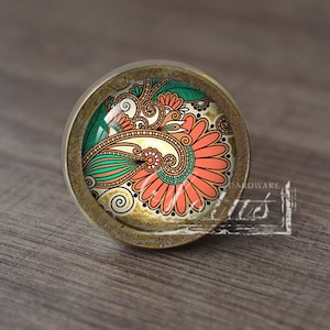 May include: A round cabinet knob with a floral design. The knob has a bronze-coloured metal base and a glass insert with a colourful floral pattern in shades of orange, green, and cream. The design is intricate and detailed.