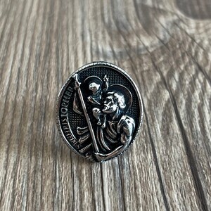 Op de afbeelding: Een zilveren ring met een gedetailleerd beeld van Sint Christoffel die een kind draagt. De ring is gegraveerd met de woorden "Saint Christopher" rond de rand.