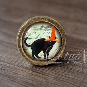 Może przedstawiać: Okrągła gałka do szafki z antycznego mosiądzu z czarnym kotem w pomarańczowym kapeluszu czarownicy. Kot jest umieszczony na tle wyblakłego pisma. Dekoracyjny element wyposażenia na Halloween.