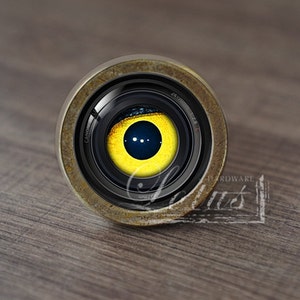 Puede incluir: Primer plano de un objetivo de cámara con un ojo amarillo en su interior. El objetivo está hecho de metal y tiene un aspecto desgastado y envejecido. El ojo está rodeado por un anillo negro y tiene un iris azul oscuro con tres puntos blancos.
