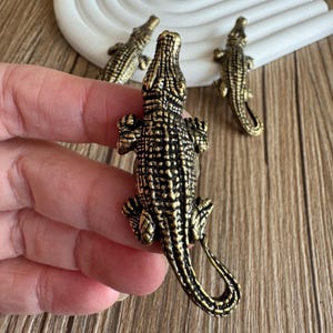 May include: Detailed bronze-colored alligator figurine with textured scales and open mouth. The alligator is held in a hand, with two other similar figurines in the background. The figurines are on a wooden surface.