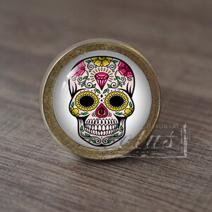 Puede incluir: Un pomo de armario redondo de color latón con un fondo blanco y un diseño de calavera de azúcar colorido. La calavera tiene flores rosas, hojas verdes, flores amarillas y un diamante en el centro de su frente.