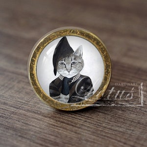 Könnte beinhalten: Ein runder, goldfarbener Schrankknopf mit weißem Hintergrund und einem Schwarzweißbild einer Katze, die einen schwarzen Hut und eine Perlenkette trägt.
