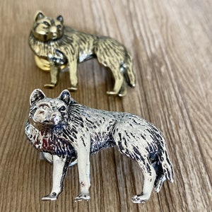 Può includere: Due figurine di lupo in metallo. Una è dorata, l'altra argentata. Entrambe sono in posizione eretta, con una texture di pelliccia dettagliata. Le figurine sono su una superficie di legno.