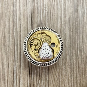 May include: A silver and gold circular pendant featuring a detailed clockwork design with a silver strawberry at its centre.