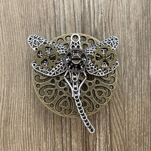 Może przedstawiać: Srebrny i brązowy wisiorek w kształcie ważki z misternymi detalami zębatych k&oacute;ł. Ważka jest umieszczona na okrągłym tle z ozdobnym wzorem.