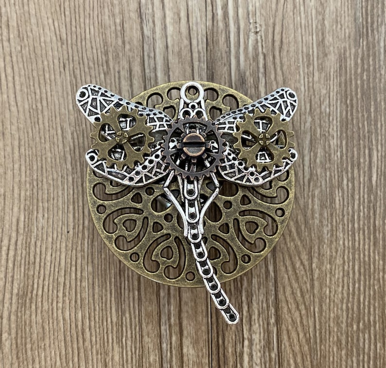 Może przedstawiać: Srebrny i brązowy wisiorek w kształcie ważki z misternymi detalami zębatych k&oacute;ł. Ważka jest umieszczona na okrągłym tle z ozdobnym wzorem.