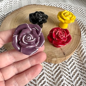 Puede incluir: Tres pomos de cajón de cerámica con forma de rosas en flor en colores morado, negro, amarillo y rojo. Cada pomo presenta un acabado brillante con capas de pétalos detalladas, diseñados para montarse en muebles para uso decorativo.