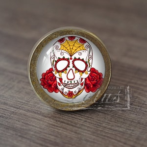 Puede incluir: Un pomo de armario redondo de color latón con un fondo blanco y un diseño de calavera de azúcar colorido. La calavera tiene detalles amarillos y rojos y está rodeada de dos rosas rojas.