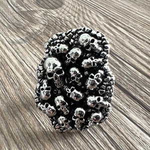 K&ouml;nnte beinhalten: Eine silberfarbene, rechteckige G&uuml;rtelschnalle, dicht bedeckt mit kleinen Sch&auml;delformen. Die Sch&auml;del sind aufwendig detailliert, mit sichtbaren Augenh&ouml;hlen, Nasen und Z&auml;hnen. Die Schnalle hat einen dunklen, strukturierten Hintergrund, der einen auff&auml;lligen Kontrast erzeugt.