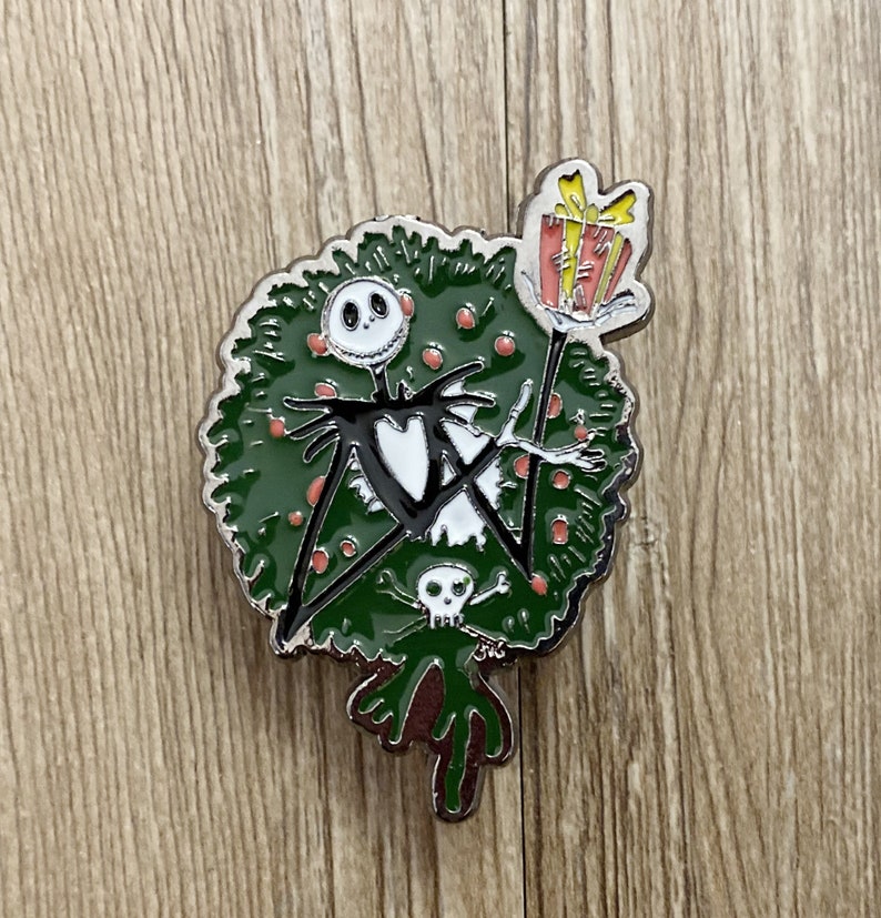 以下が含まれることがあります： ナイトメア・ビフォア・クリスマスのジャック・スケリントンを描いた緑と赤のエナメルピン。彼はプレゼントを持って、クリスマスリースの前に立っています。