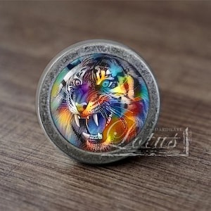Puede incluir: Un pomo de armario redondo de color plateado con la cara de un tigre colorido. Los ojos del tigre son azules y verdes, y su boca está abierta en un rugido.