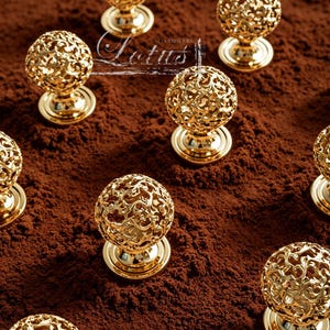 May include: Gold-colored cabinet knobs with an ornate, openwork sphere design. The knobs sit on a round base and are arranged on a textured, brown surface. The image includes the text "Lotus Hardware".