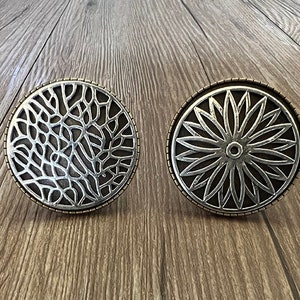 May include: Two silver metal decorative drawer pulls with intricate designs. One pull features a floral pattern, while the other has a more organic, web-like design.