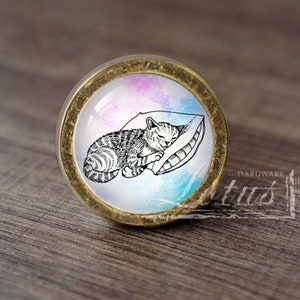 May include: A round, gold-colored cabinet knob with a white background and a black and white illustration of a cat sleeping on a pillow. The background has a watercolor effect with pink, blue, and purple hues.