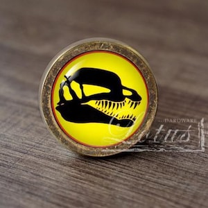 May include: A round cabinet knob with a bronze-toned metal frame and a glass insert. The insert features a bright yellow background with a black silhouette of a dinosaur skull facing right, enclosed by a thin red circular border.