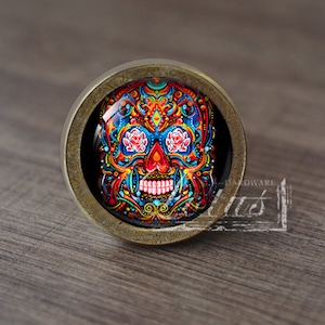 Puede incluir: Un pomo de armario redondo de color latón con un diseño de calavera de azúcar colorido y detallado en la parte delantera. La calavera está rodeada de un fondo negro.