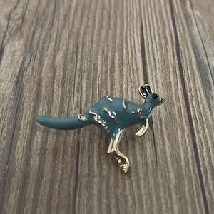 Könnte beinhalten: Graue Känguru-Brosche mit goldfarbenen Akzenten. Die Brosche aus Metall hat eine glänzende Oberfläche. Das Känguru ist in einer springenden Pose dargestellt, mit ausgestrecktem Schwanz. Ein dekoratives Accessoire.