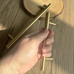May include: Gold-colored cabinet pulls with a T-bar design. The pulls are cylindrical with a perpendicular bar at each end. One pull is held in a hand, while two others are on a wooden surface.