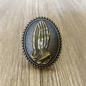 May include: A gold-toned metal knob with a raised design of a praying hand. The knob has a rope-like border around the edge.