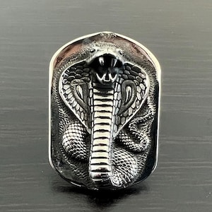 May include: A silver-toned pendant featuring a detailed, three-dimensional sculpture of a cobra with its hood raised. The cobra's scales and eyes are intricately sculpted.