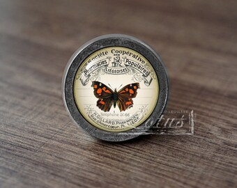 May include: A round, silver knob with a vintage-style label featuring a butterfly and the text "Pharmacies Populaires Société Coopérative Liegeoises Telephone 30.66 H.GILLARD PHARMACIEN Rue d'amercœur. 71. LIEGE".