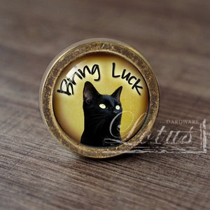 Peut inclure: Bouton de meuble rond en laiton vieilli avec une illustration de chat noir sur fond jaune. Le texte "Bring Luck" est écrit au-dessus du chat. Le bouton est posé sur une surface en bois.
