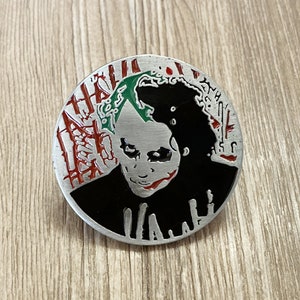May include: A silver metal pin with a black and green image of the Joker from Batman. The Joker's face is painted with a white smile and red lipstick. The background is a red and white pattern.