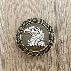 May include: A silver eagle head with a detailed design is featured on a round, bronze-coloured metal pin with a decorative border.